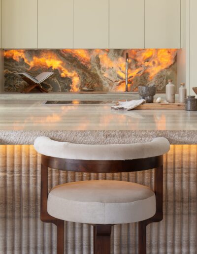 Modern kitchen with a marble island, three cushioned bar stools, a vase of white tulips, and a unique orange stone backsplash with under-cabinet lighting.